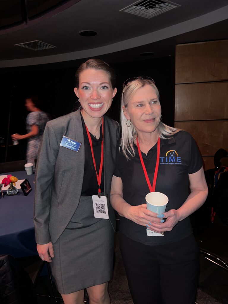 Gillain Keller and Kate Rounding stand indoors, smiling. One wears a gray blazer and skirt with a red lanyard and name badge; the other wears a black polo shirt with a TIME logo, holding a cup and also wearing a red lanyard.