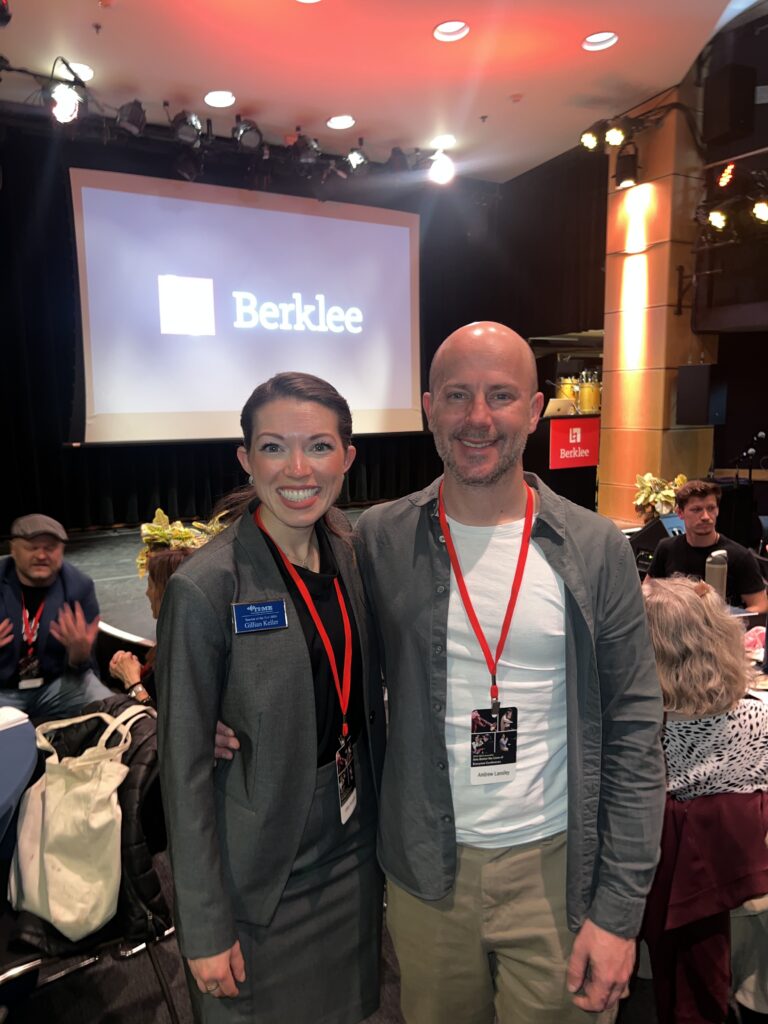 Gillian Keeler and Andrew Landsley wearing event lanyards smile and pose together in a room with tables and people. A large screen behind them displays the Berklee logo. The atmosphere is lively, with stage lights and decorations visible.