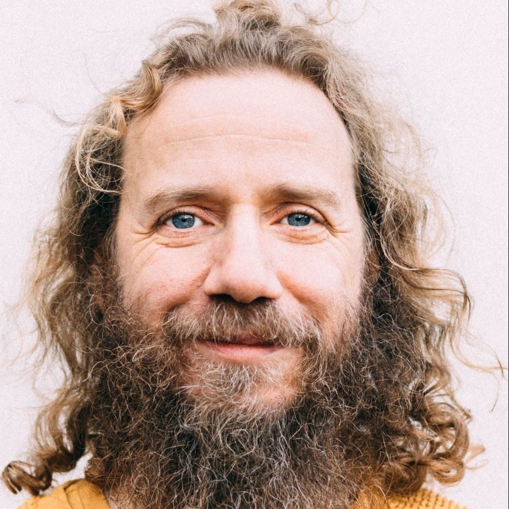 A person with long curly hair and a full beard smiles gently at the camera, wearing a mustard yellow sweater. The background is plain and light-colored.