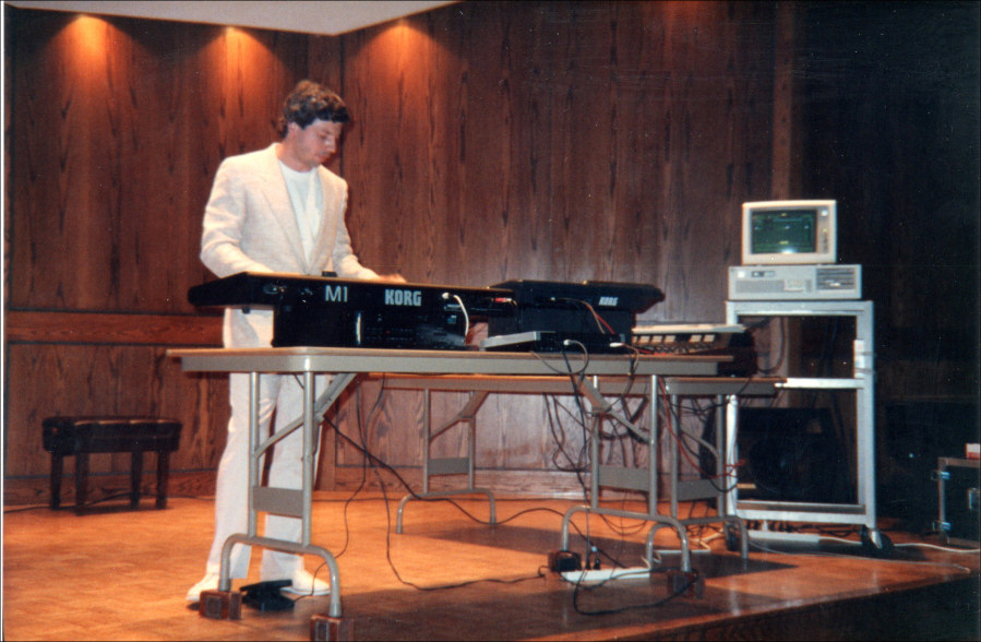 A man in a white suit plays a Korg keyboard onstage, with audio equipment and a vintage computer setup on a cart beside him. The background is wood-paneled walls.