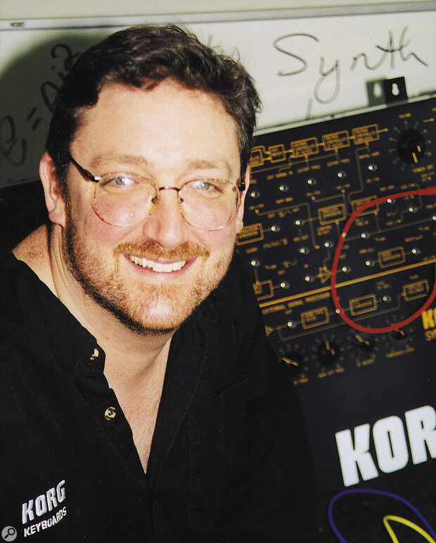 A smiling man with glasses and a beard stands in front of a Korg synthesizer, wearing a black Korg shirt. The background includes handwritten text on a whiteboard and close-up details of the synthesizer.