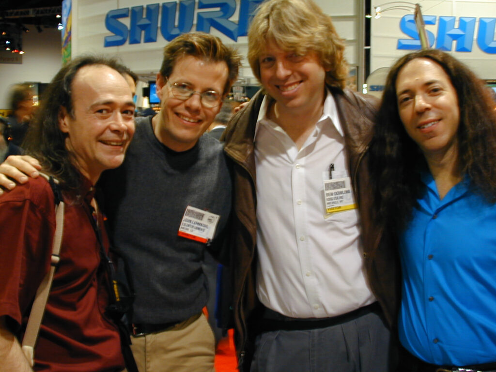 Four men stand together indoors, smiling at the camera. They are at an event or convention, wearing name badges, with SHURE signage visible in the background.
