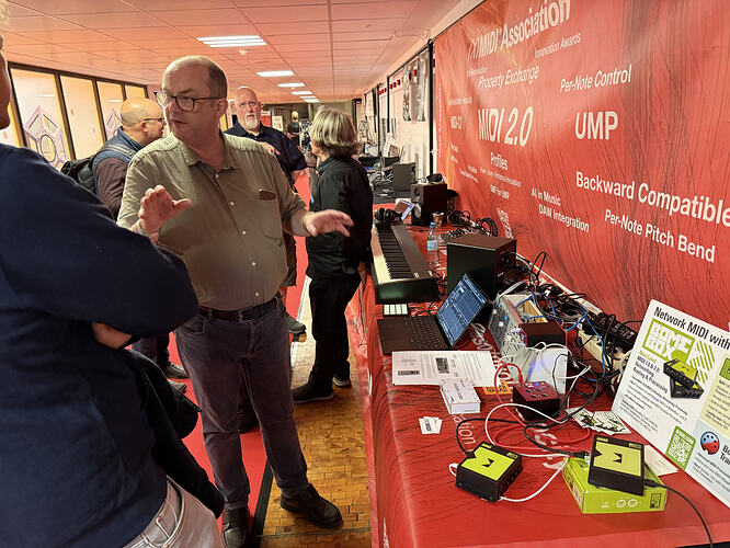 A man gestures while talking to another person at a tech expo table displaying various MIDI devices, cables, and electronic equipment, with a large red MIDI Association banner in the background.