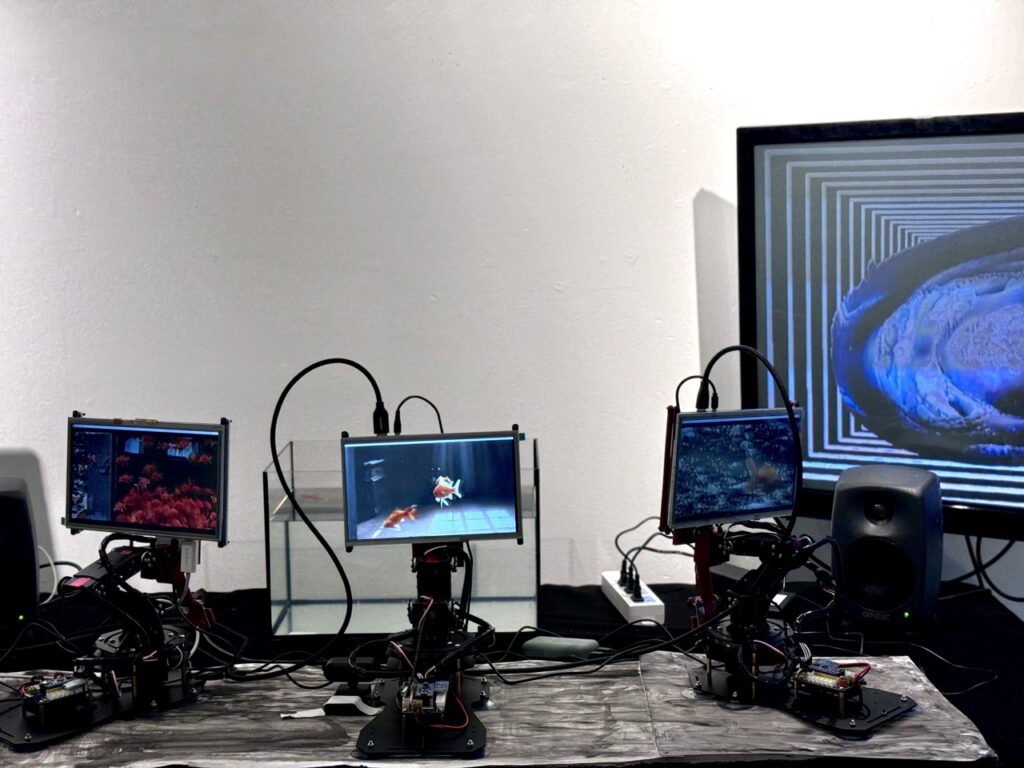 Three small screens on robotic arms display underwater scenes, positioned on a table with cables and electronics. In the background, a large monitor shows a spiral visual with blue tones, next to a speaker and an empty glass tank.