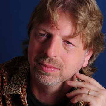 A middle-aged man with wavy light brown hair, short beard, and mustache poses against a blue background. He is wearing a patterned shirt and gently touching his face with one hand, displaying a gold ring.