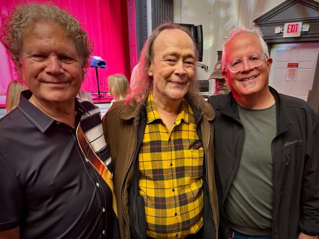 Three smiling older men stand close together indoors, posing for a photo. The man in the middle wears a yellow plaid shirt and brown jacket, while the other two wear glasses and dark jackets. A red curtain is in the background.