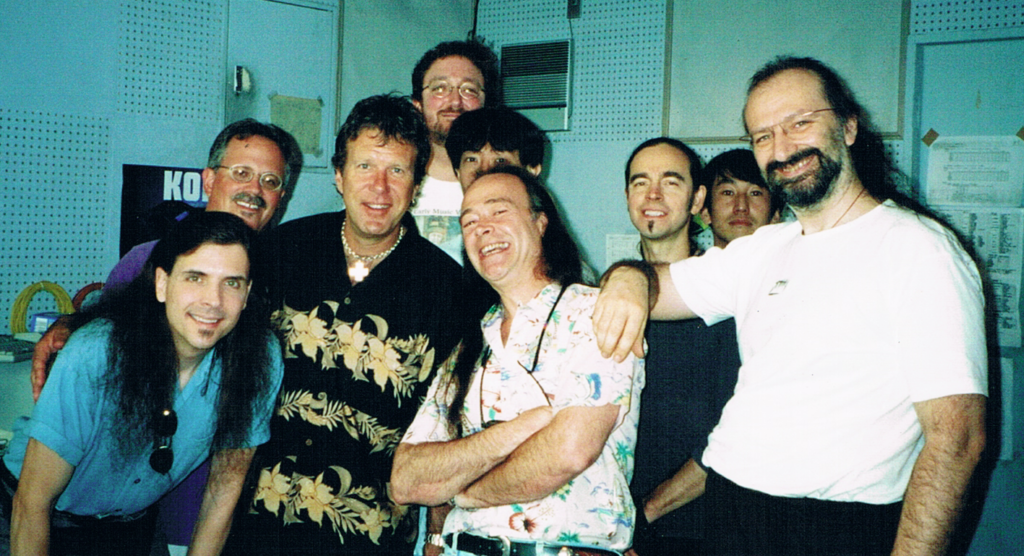 A group of nine men, some smiling and some with neutral expressions, stand together indoors in a casual setting. Most have long hair, and they are dressed in casual shirts. The background shows a blue wall and some posters.