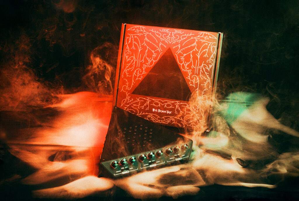 A dramatic, smoky scene features a mysterious box with ornate, golden drawings and a triangle cut-out, alongside a sound mixer with knobs and lights. Red and green lighting adds an eerie atmosphere.