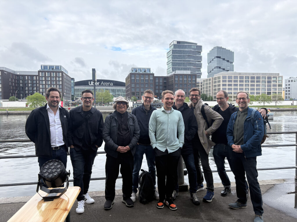 MIDI Association Members from Steinberg, Native Instruments, Music Hackspace and more stand smiling together outdoors near a waterfront, with modern office buildings and the Uber Arena visible in the background under a cloudy sky.
