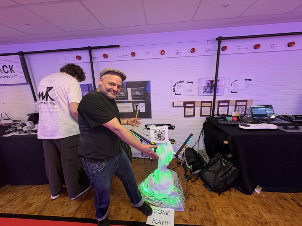 A Toni Hunke from Abletondrummer.com stands in front of an interactive ConnectThroughMIDI exhibit, pointing at a display with a green light and a COME PLAY!! sign. Another person is adjusting equipment at a nearby table with electronics and posters.