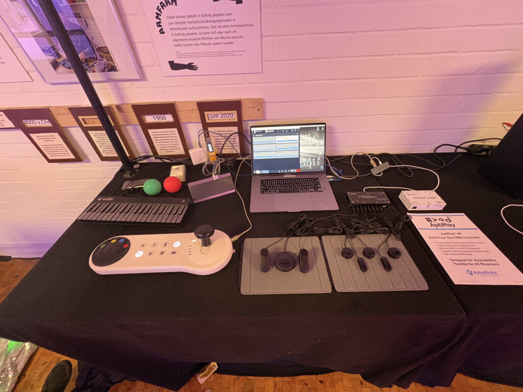 An accessible music technology display on a table, featuring adaptive controllers, various buttons, a laptop with music software, and informational signs, designed for inclusive music-making.