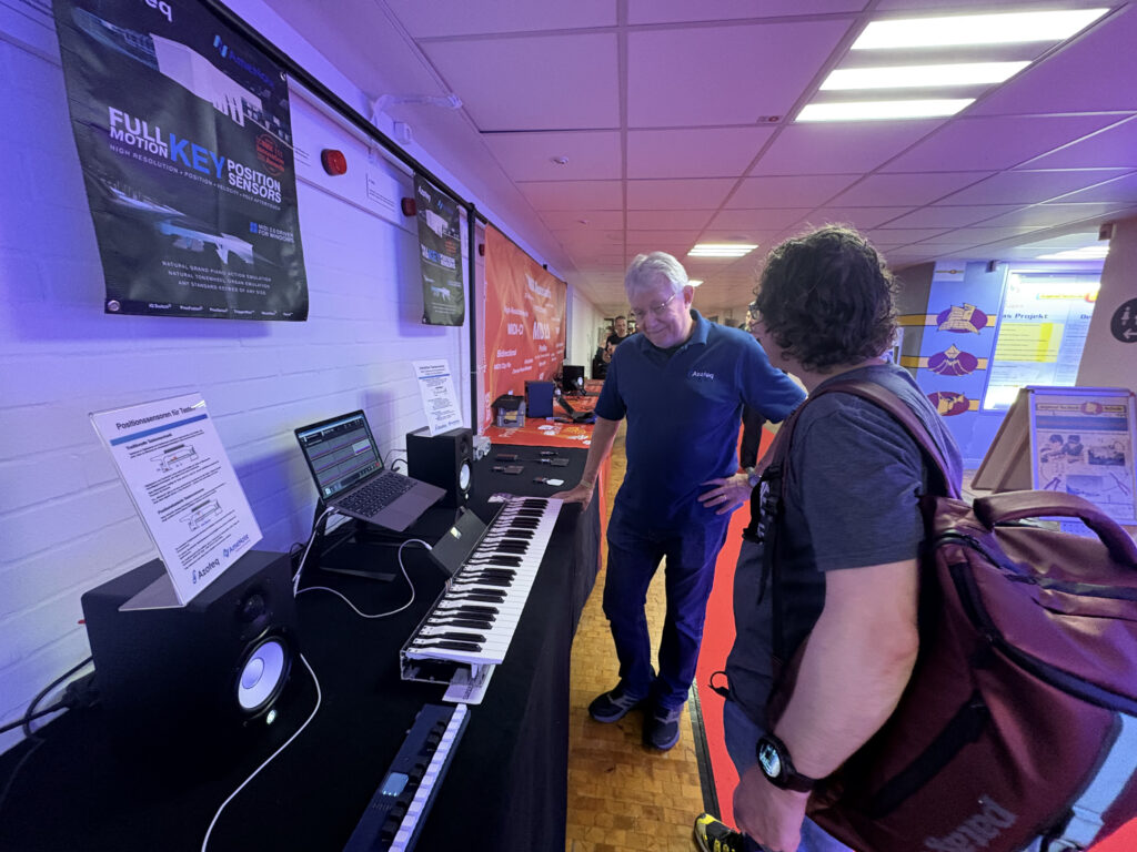 Two people stand by a table displaying a MIDI keyboard, laptop, and speakers at an indoor event or exhibition. Posters and information sheets are on the wall and table. Blue and purple lighting fills the space.