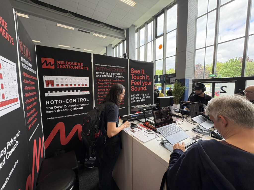 A group of people interact with music production equipment at a Melbourne Instruments booth, featuring banners and demo stations, in a well-lit room with large windows and green trees visible outside.
