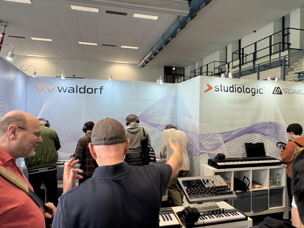 People testing electronic musical instruments and synthesizers at a booth featuring Waldorf, Studiologic, and Sonic logos in a trade show or exhibition. A man in the foreground is gesturing toward the display.