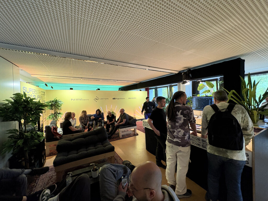 A group of people gather in a modern lounge with sofas and plants, engaged in conversation and activities. Several people sit, while others stand around a table with electronic equipment near a green-lit wall.