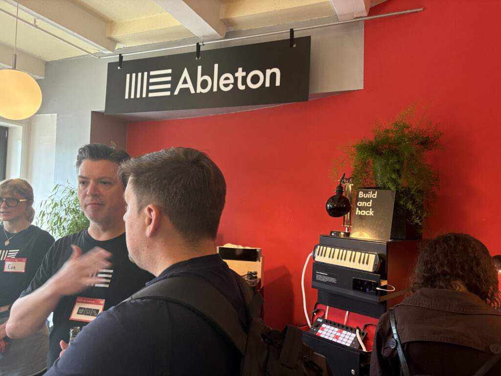 People gather and talk in front of an Ableton sign on a red wall. Music equipment and a “Build and hack” sign are displayed on a table next to a green plant. Name tags are visible on some attendees.