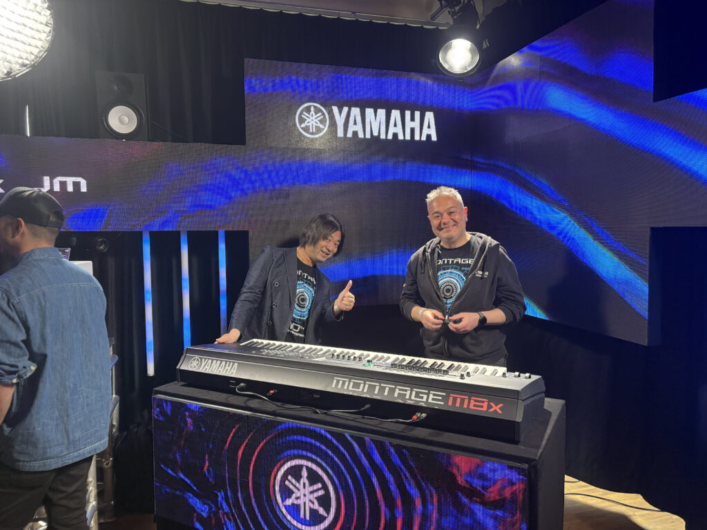 Two men stand behind a Yamaha MONTAGE M8x keyboard on a stage with Yamaha branding. One man smiles and gives a thumbs up, while the other also smiles. The background features blue lighting and abstract patterns.