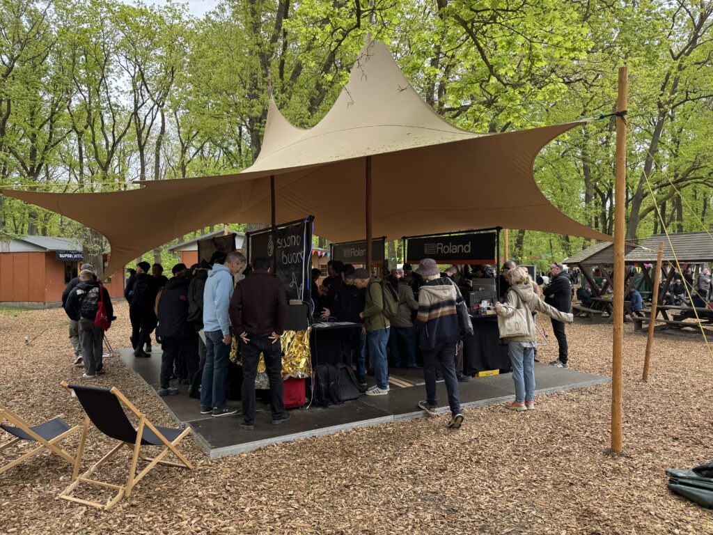 A group of people gather under a large tan canopy tent in a wooded outdoor area, interacting with exhibitors at display tables. Surrounding trees are lush and green; some empty lounge chairs sit nearby.
