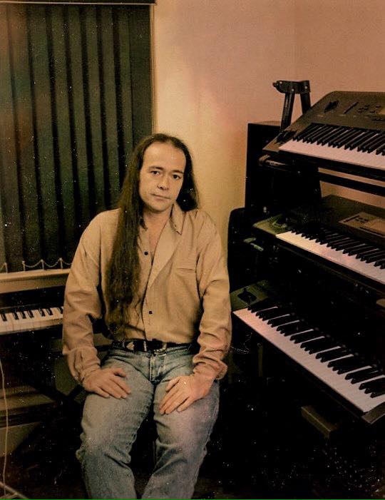 Jack Hotop with long hair, wearing a beige shirt and jeans, sits on a stool beside several stacked electronic keyboards and a piano in a dimly lit room with closed blinds.