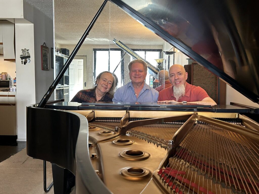 Three smiling men sit closely together behind a grand piano, with the open strings and interior of the piano visible in the foreground. The setting appears to be a cozy, well-lit living room.