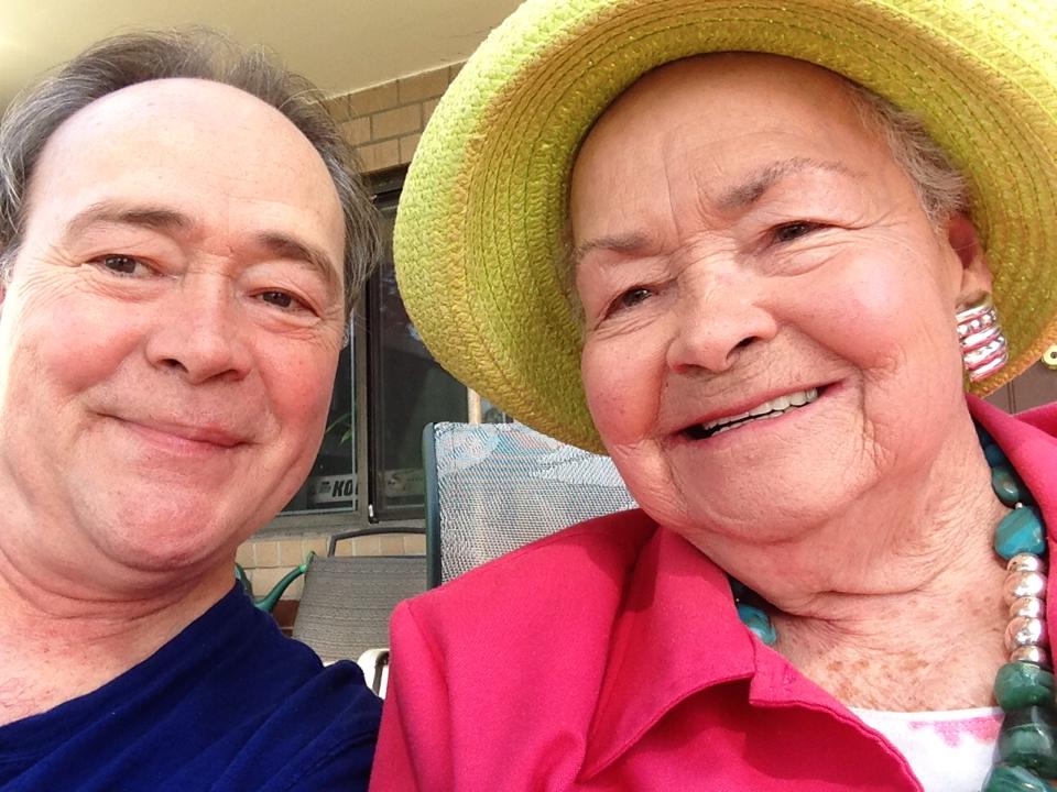 A smiling man and an older woman in a straw hat and pink jacket sit close together outdoors, posing for a cheerful selfie.