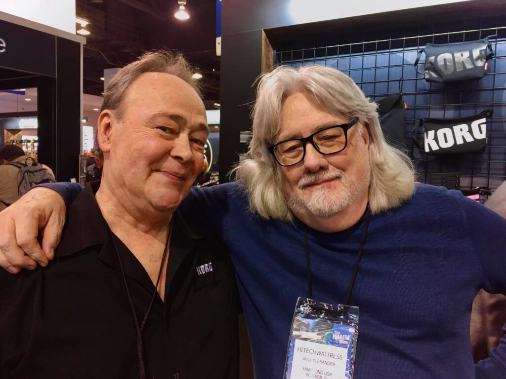 Jack and Keith Emerson keyboard Tech will Alexander pose together at an event, one wearing a black Korg shirt and the other in a blue sweater with glasses and a badge. Korg-branded gear is displayed in the background.