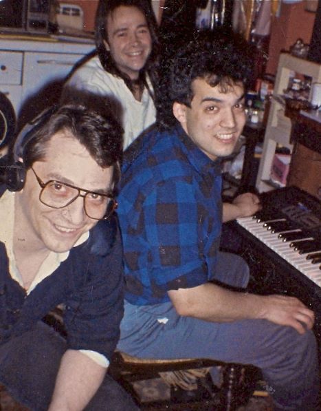 Three men indoors, smiling at the camera; one wears glasses and headphones, another sits at a keyboard, and the third is in the background, all appearing relaxed and happy in a casual setting.