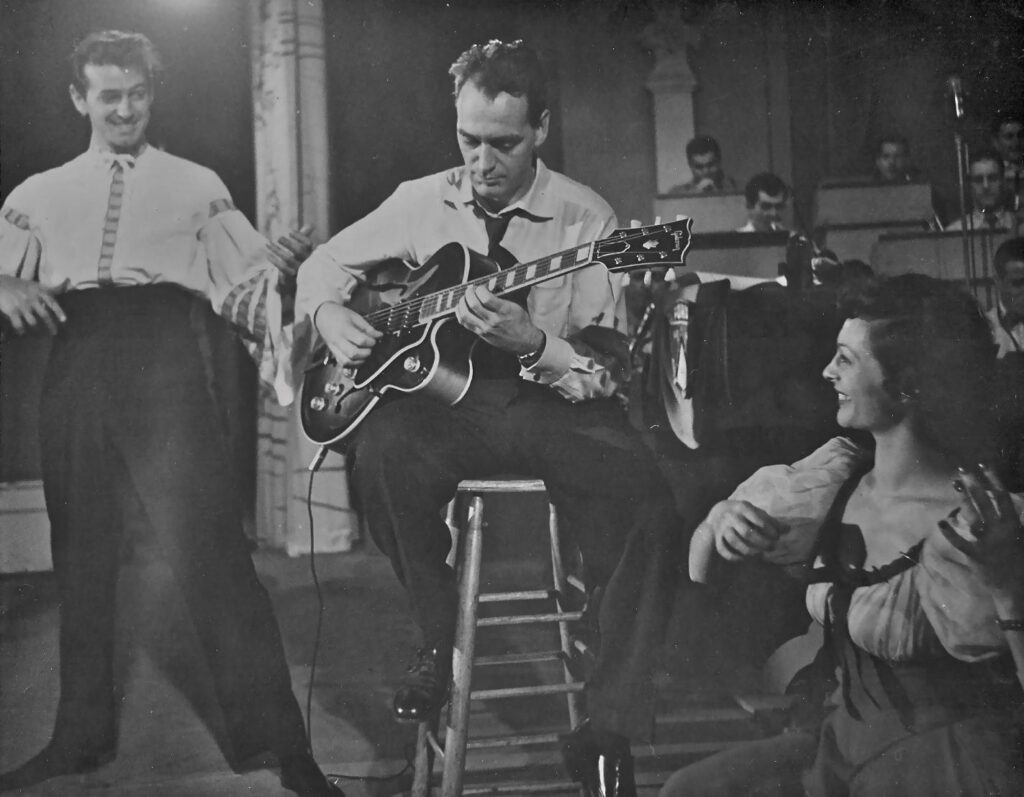 Jack Hotop Sr sits on a stool playing an electric guitar while another man and a smiling woman, both in costume, watch him. An orchestra is visible in the background. The scene appears to be from a performance or rehearsal.