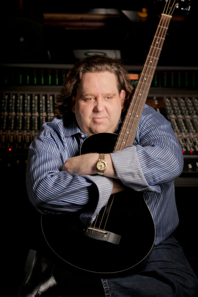 A man with medium-length brown hair sits in front of an audio mixing console, wearing a blue striped shirt and a gold watch, and holds a black acoustic guitar across his chest.