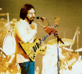 A musician stands on stage playing a bass guitar and singing into a microphone, with drums and other instruments visible in the background under stage lighting.