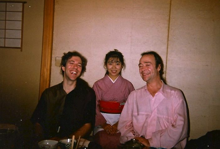 Three people sit together indoors, smiling for the camera. The woman in the center wears a pink kimono with a red obi, while the two men on either side wear casual shirts. Dishes and cups are visible on the table in front of them.
