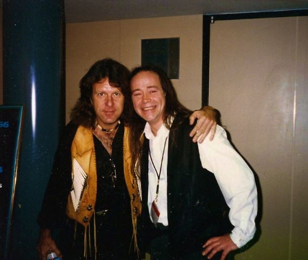 Keith Emerson and Jack people are standing together indoors, smiling at the camera. The person on the left has long hair and wears a tan vest over a black shirt. The person on the right also has long hair and wears a white shirt with a black vest.