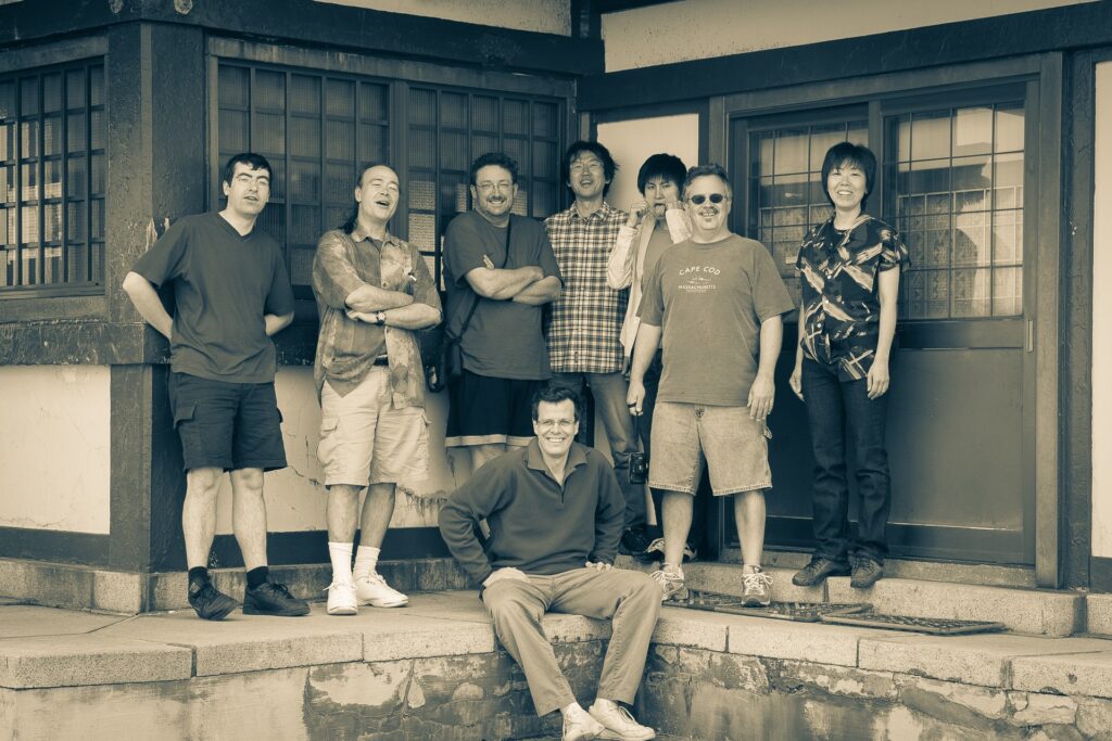 A group of eight people, seven standing and one sitting on a step, pose and smile outside a building with barred windows and wooden beams in a casual, sepia-toned photo.