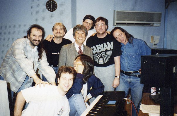 A group of eight smiling adults pose together in a recording studio, some standing and some sitting around a keyboard, with sound equipment and a wall clock visible in the background.