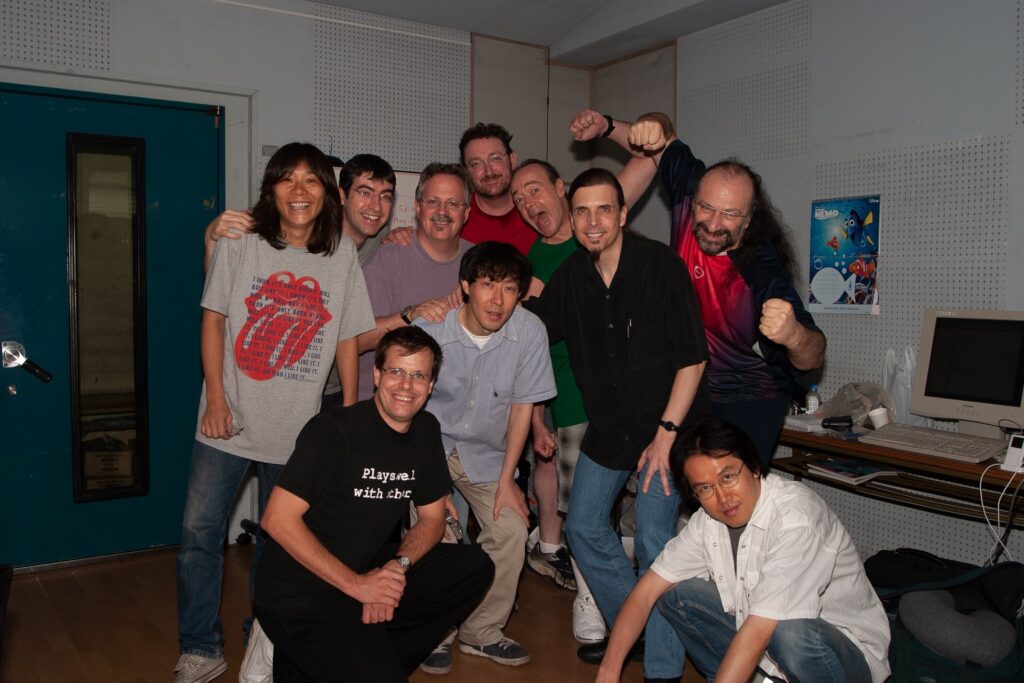 A group of ten people pose together in a recording studio, some smiling and some making playful gestures. There are posters, a desk with a computer, and soundproof walls in the background.