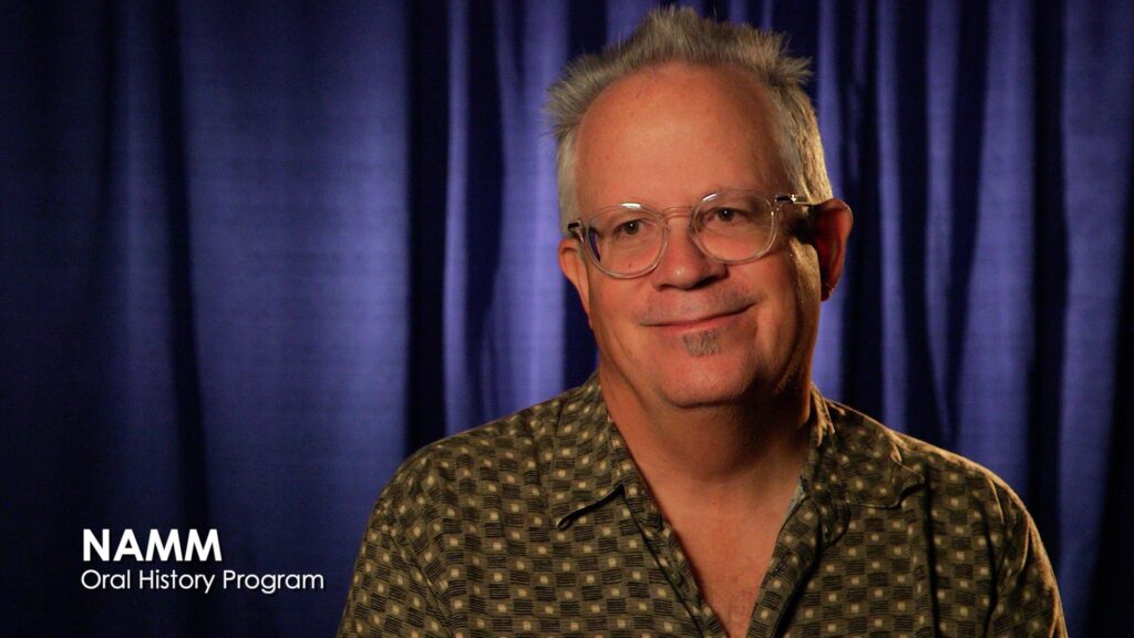 A man with short gray hair, glasses, and a patterned shirt smiles in front of a dark blue curtain. NAMM Oral History Program text is in the lower left corner.