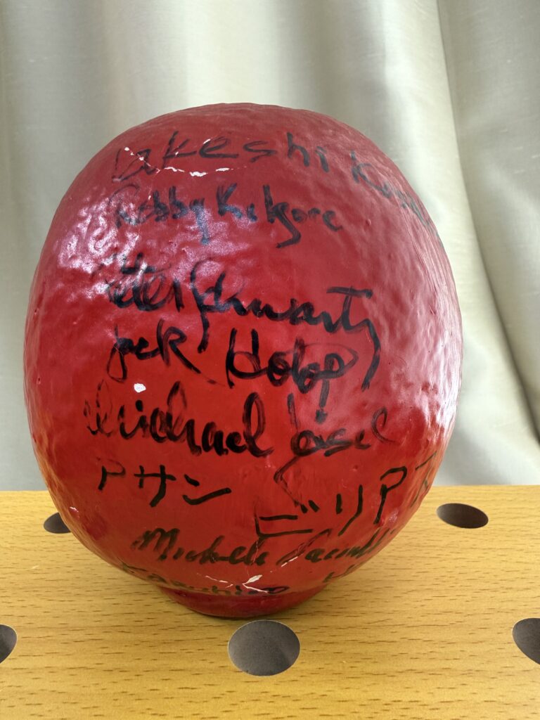 A red, spherical object covered in handwritten signatures and messages in black ink is displayed on a light brown, perforated wooden surface with a light-colored curtain in the background.