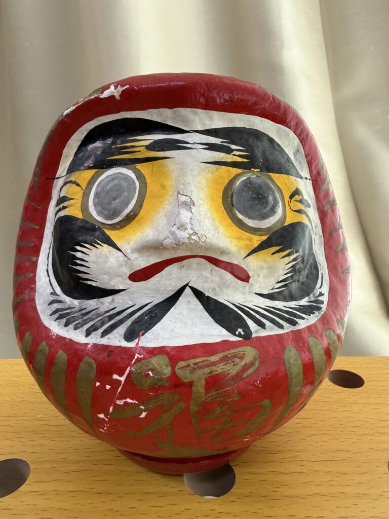 A red Daruma doll with bold black and yellow facial features, white blank eyes, and a mustache, sits on a wooden surface against a light-colored curtain background.