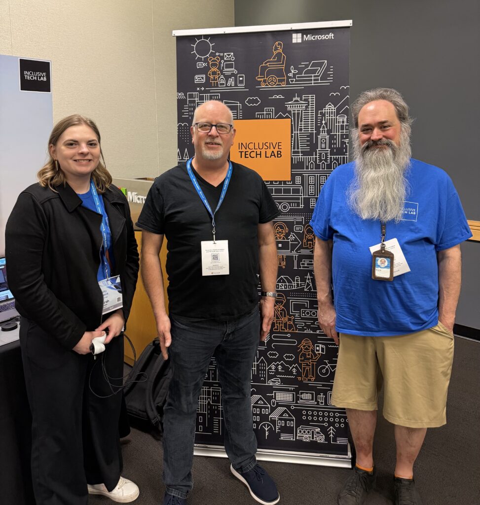 Three people standing and smiling in front of a Microsoft Inclusive Tech Lab banner. Two men (one with a long grey beard, one with glasses) and one woman, all wearing conference badges.