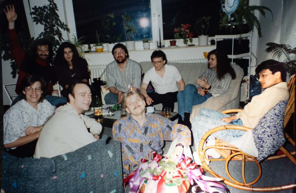 A group of nine adults sit together in a cozy living room, smiling and talking. There are plants on the windowsill behind them and drinks on the table. One person at the back waves at the camera.