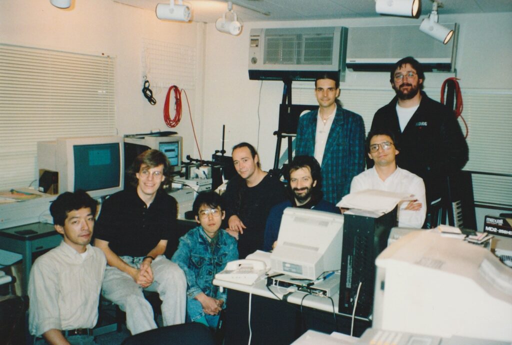 A group of eight people posing and smiling in a cluttered office filled with computers, monitors, and technical equipment. The workspace has cables, paperwork, and various electronic devices visible.