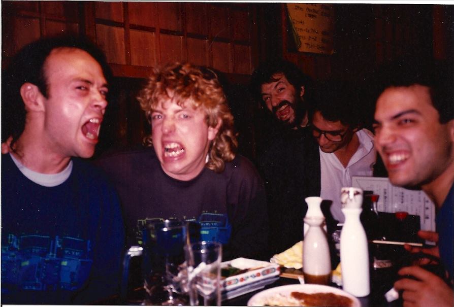 Five people sit at a table in a restaurant, laughing and making silly faces. The table is set with plates, glasses, and bottles. The background features wooden paneling. The mood appears lively and cheerful.