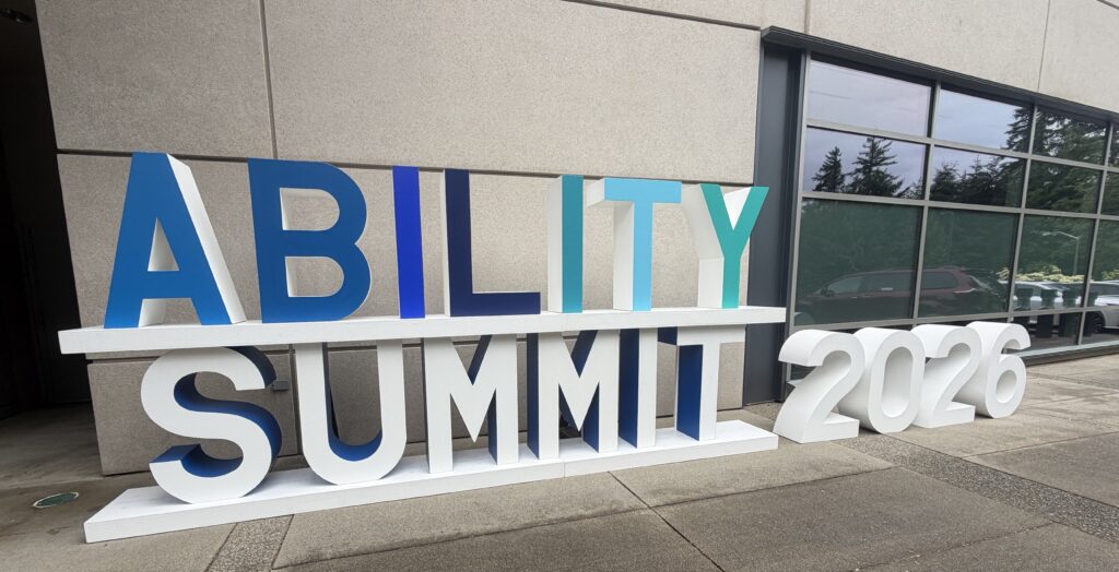 Large 3D letters spelling ABILITY SUMMIT 2026 are displayed outside a modern building with large windows, trees reflected in the glass, and a sidewalk in the foreground.