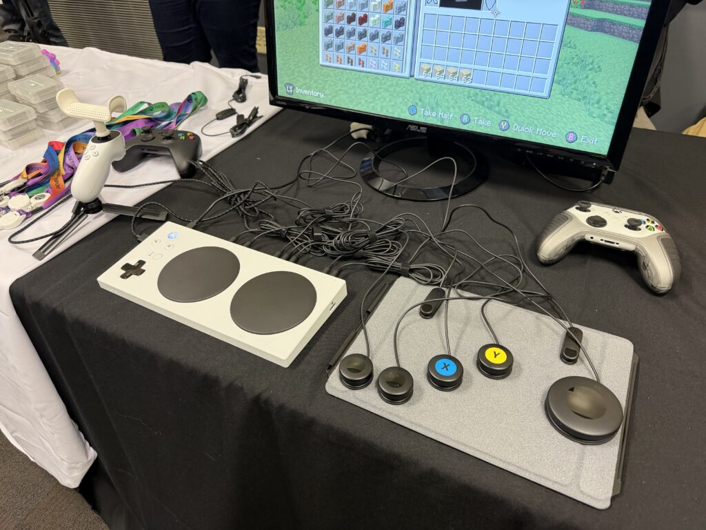 A gaming setup featuring an Xbox Adaptive Controller with large buttons and switches connected, alongside standard controllers and a monitor displaying the game Minecraft. The devices are on a black tablecloth.