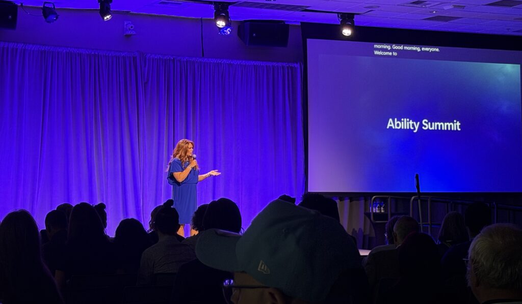 A woman in a blue dress speaks on stage at an event, with an audience seated in front. A large screen beside her displays the words Ability Summit in bold text. Blue lighting illuminates the scene.