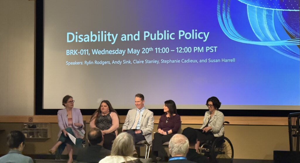 Five panelists sit in front of a screen displaying Disability and Public Policy and event details, including date, time, and speaker names. One panelist uses a wheelchair; an audience member appears to ask a question.