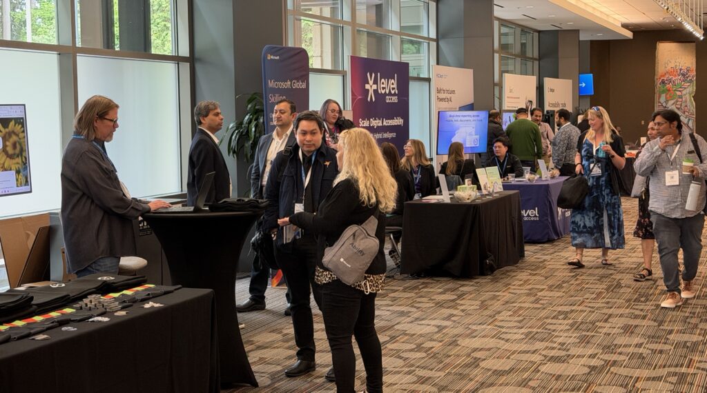 People networking and visiting booths at a professional conference or expo in a spacious, well-lit hallway with banners and tables for various companies. Attendees are engaged in conversations, and informational displays are visible.