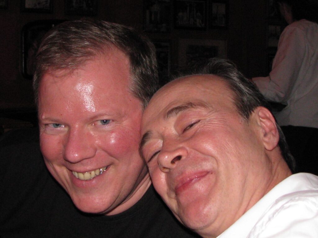 Two men are smiling closely together in a warmly lit indoor setting, appearing happy and relaxed. One man is wearing a black shirt, and the other is in a white shirt, with framed photos visible on the wall behind them.