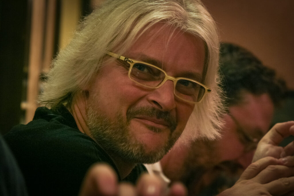 Micheal Geisel with shoulder-length gray hair, a beard, and light-colored glasses smiles at the camera in a warmly lit indoor setting. Another person is partially visible beside him.