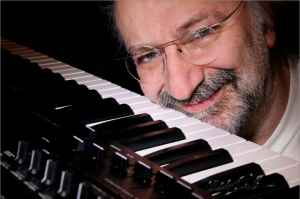 A smiling older man with glasses and a beard leans close to the keys of a synthesizer keyboard, with his face partially framed by the instrument.